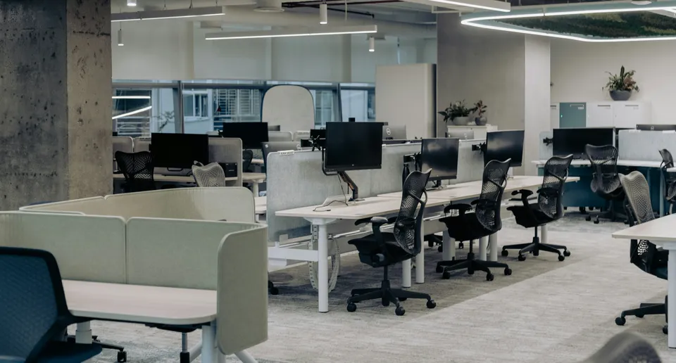 An empty open-plan office with rows of clean desks and chairs in soft daylight.