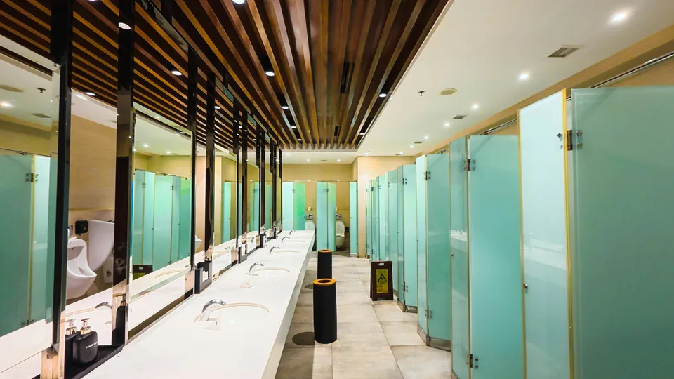 A commercial washroom with a row of sinks, mirrors and frosted glass stalls.
