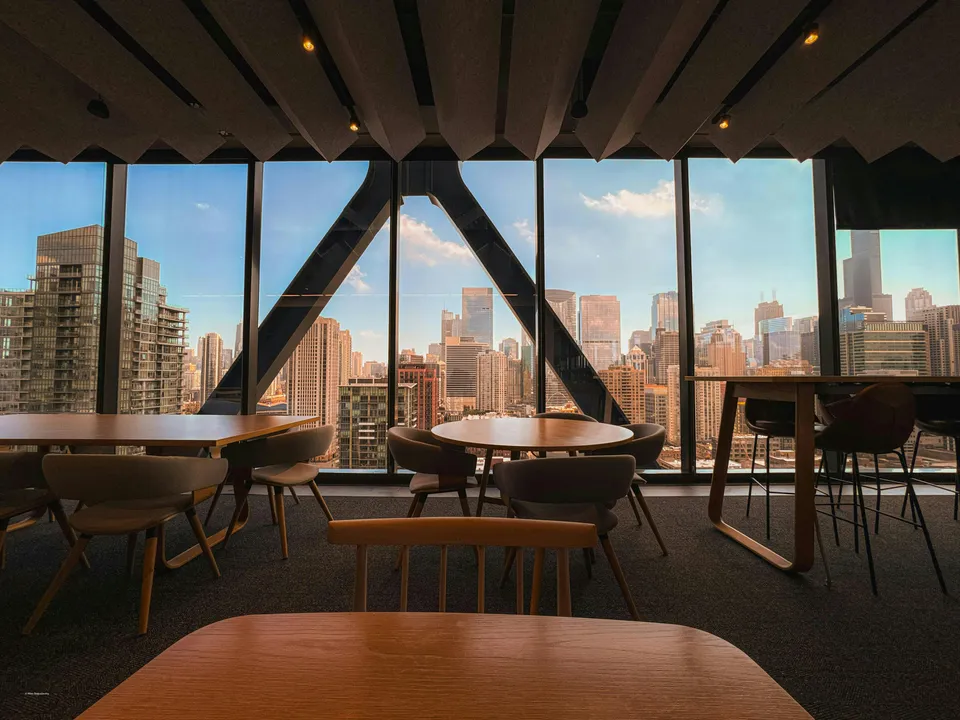 An office lounge with tables and chairs by floor-to-ceiling windows over a city skyline.